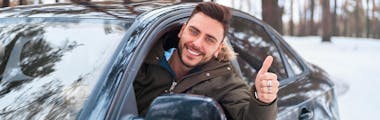 Man wearing winter coat giving thumbs up and smiling out the window in a snowy landscape