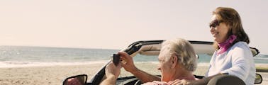 Elderly couple on a road trip in a convertible with the top down taking pictures of the ocean in the summer sun